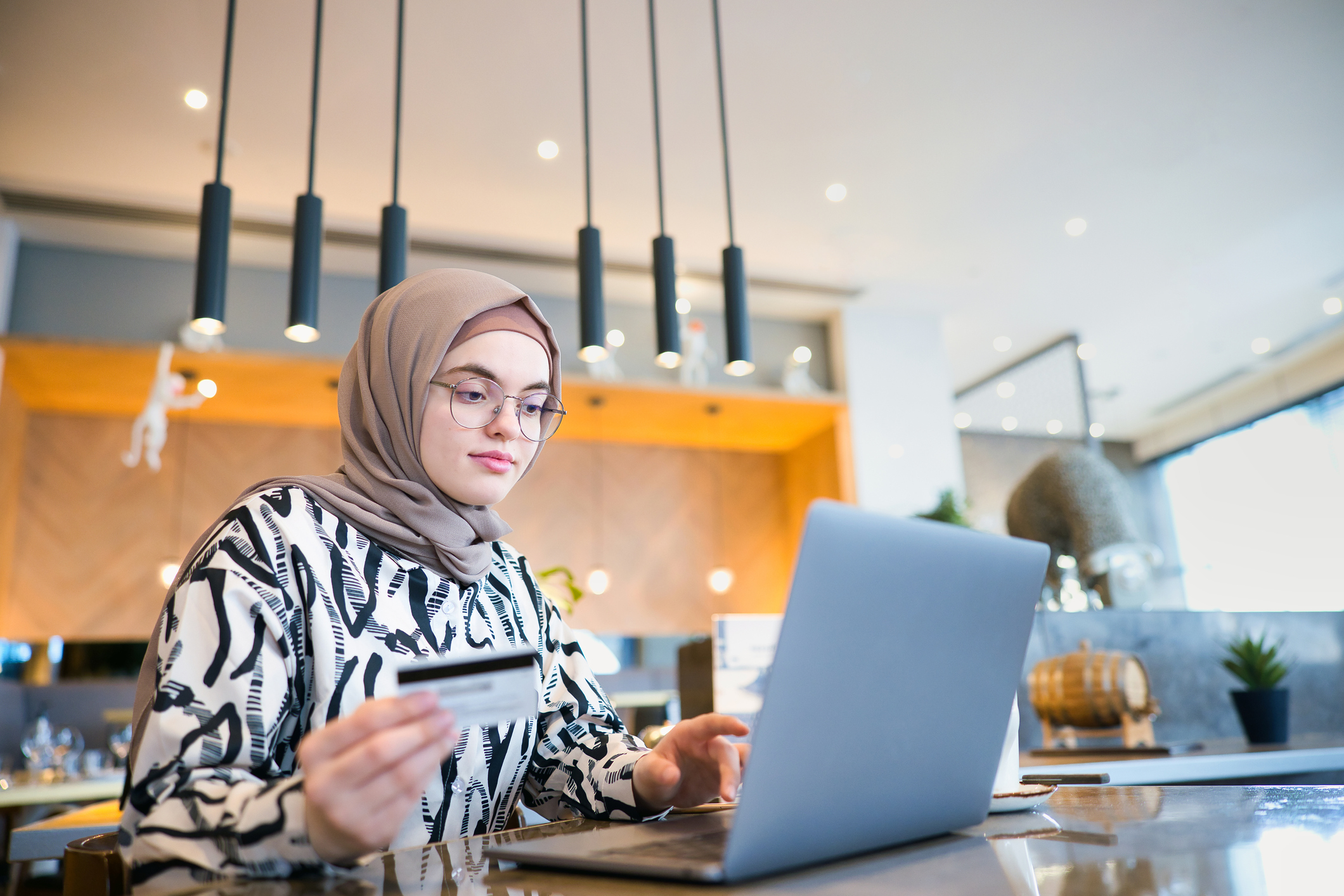 Young Arab American woman wearing a hijab scarf sits at a table in a cafe. She is holding her credit card in her right hand, and has her laptop open to pay a monthly student bill online.