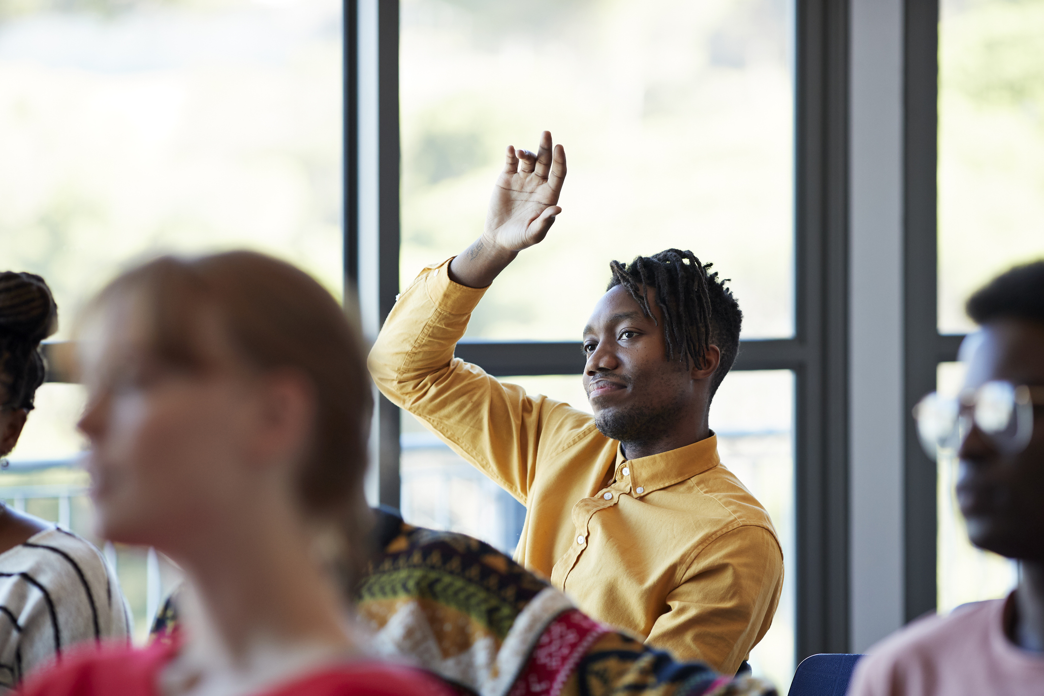 Black male community college student sitting at a desk in a classroom with other students. His arm is raised to answer a question from the professor off-screen.