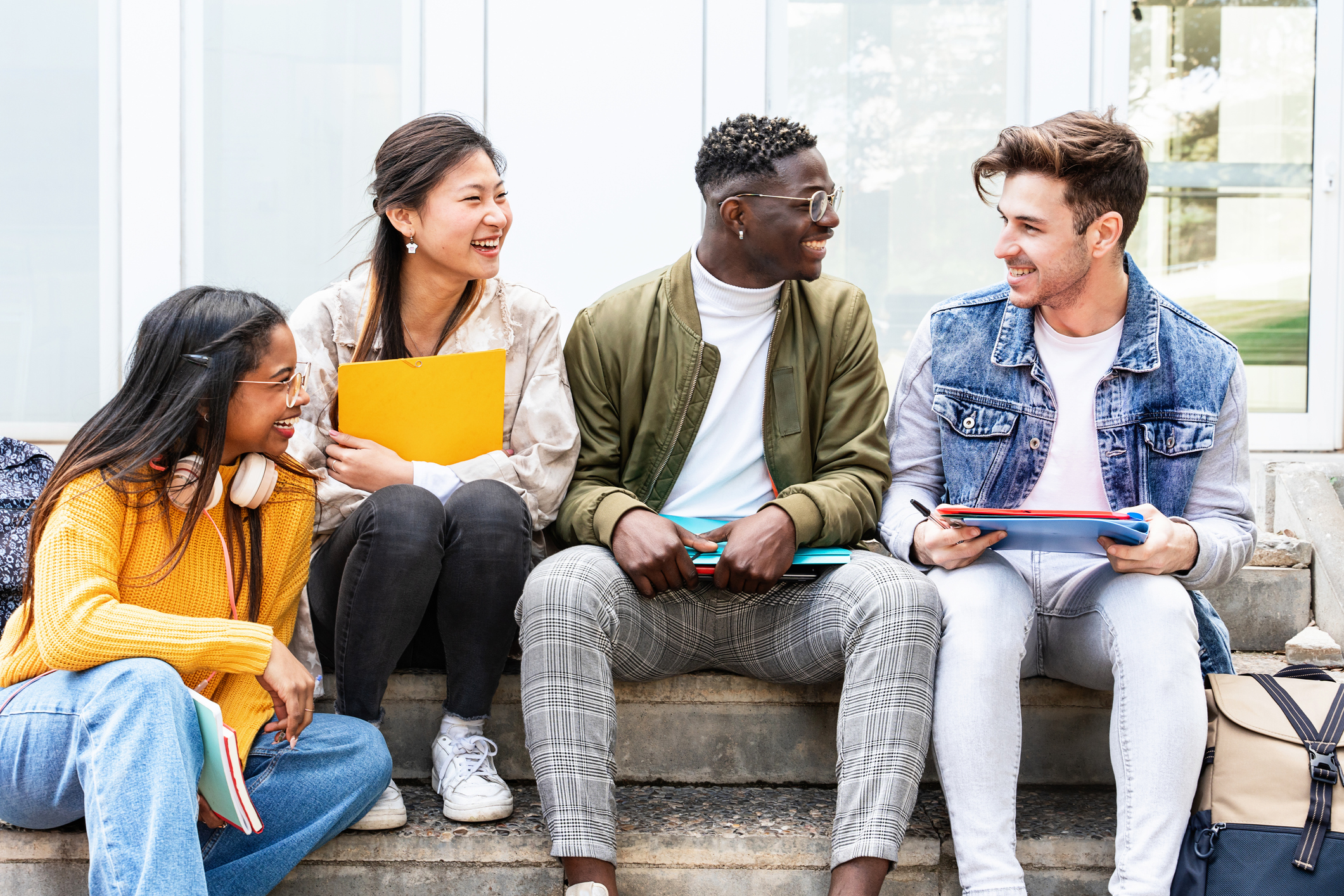 A multicultural group of four friends sitting on steps outside of a university campus building. They are laughing and chatting between classes.