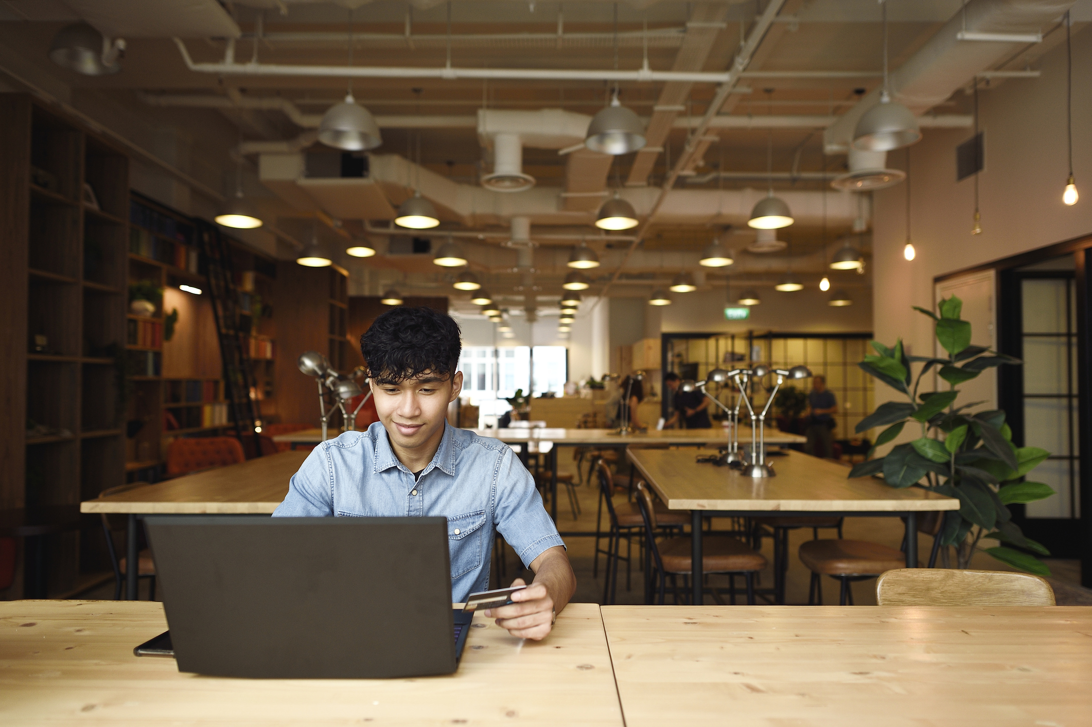 A male Asian American college student sitting at a table in a student center. He has his laptop open, and is using his credit card to pay for a school tuition bill online.