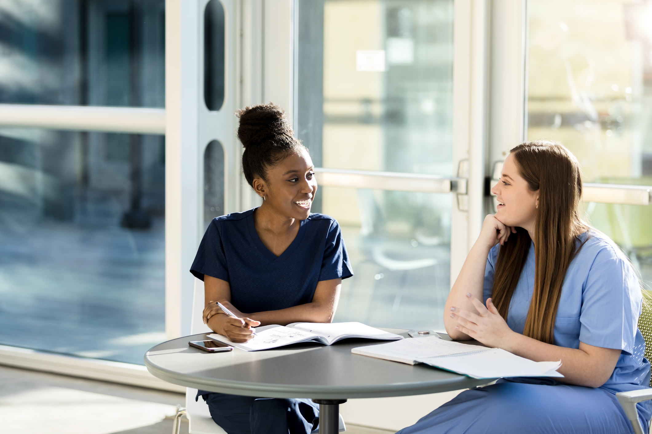 Two female medical students wearing blue scrubs sit at a table in a classroom building lobby. They are chatting and reading over their notes while studying for an exam.