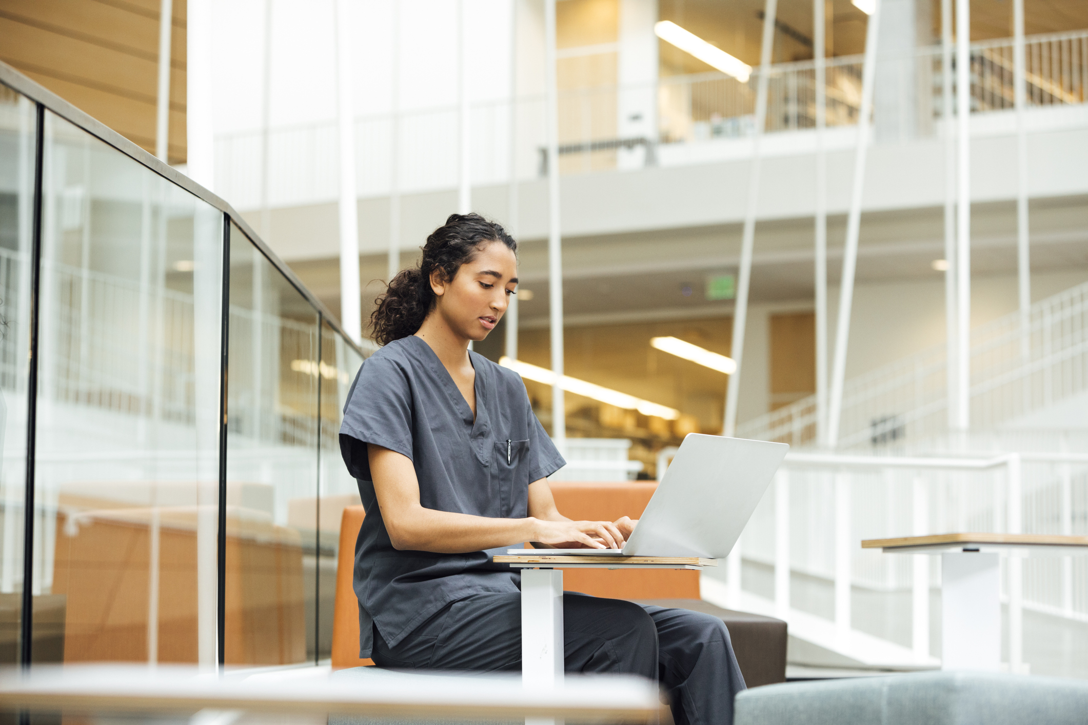 Female Middle Eastern medical student sitting at a table in a university building atrium. She is typing on her laptop while studying for her MCAT.