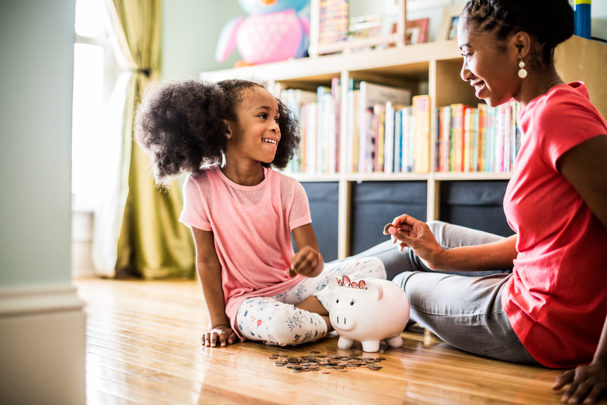 A Black mother and her young daughter are sitting on the floor in the living room at home. The mother is teaching her daughter about saving money as she helps count coins in her daughter's piggybank.