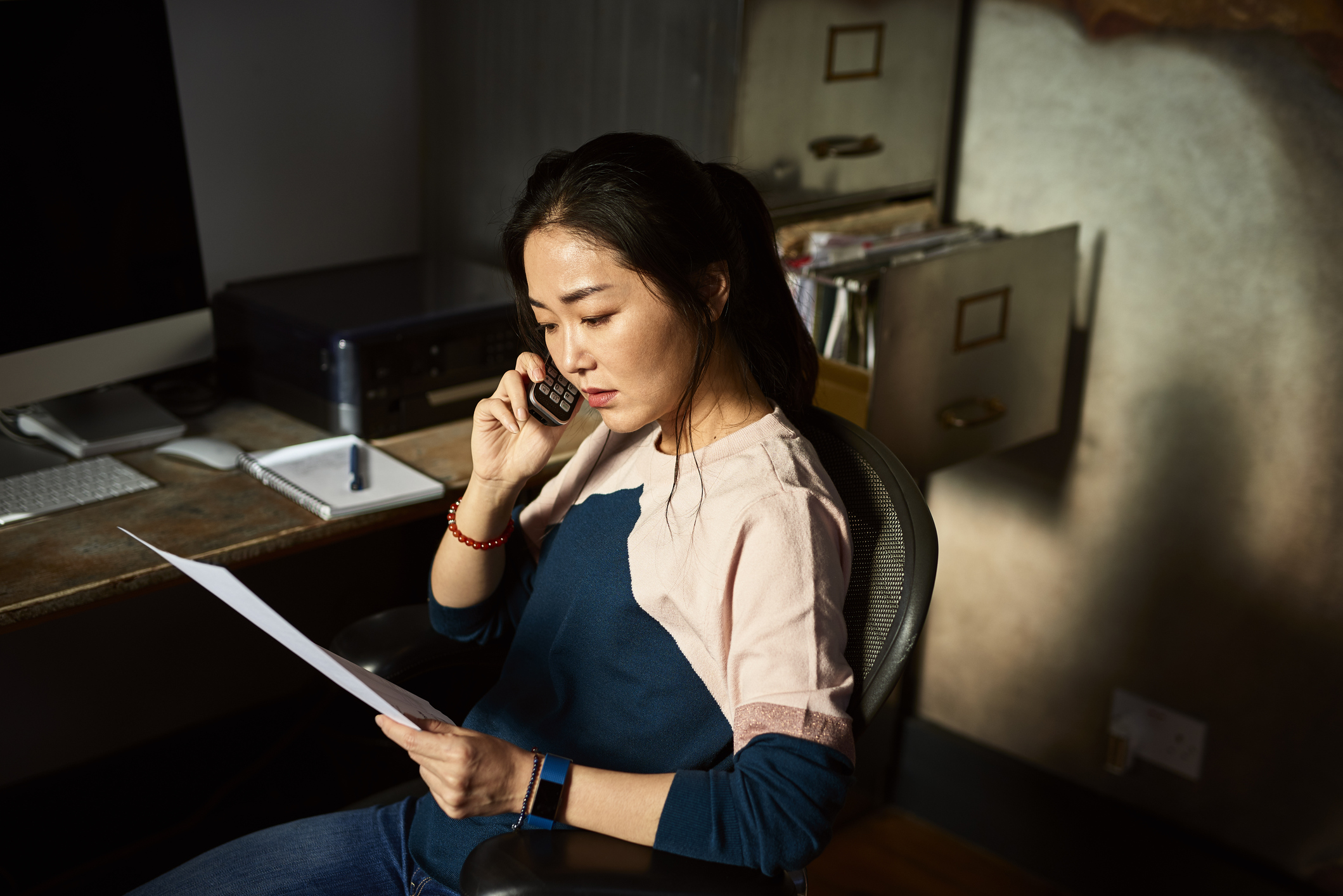 Serious mid adult Asian woman listening on phone, sitting in office chair in dark room, holding paperwork, checking accounts, financial planning, money concerns, difficulty, debt