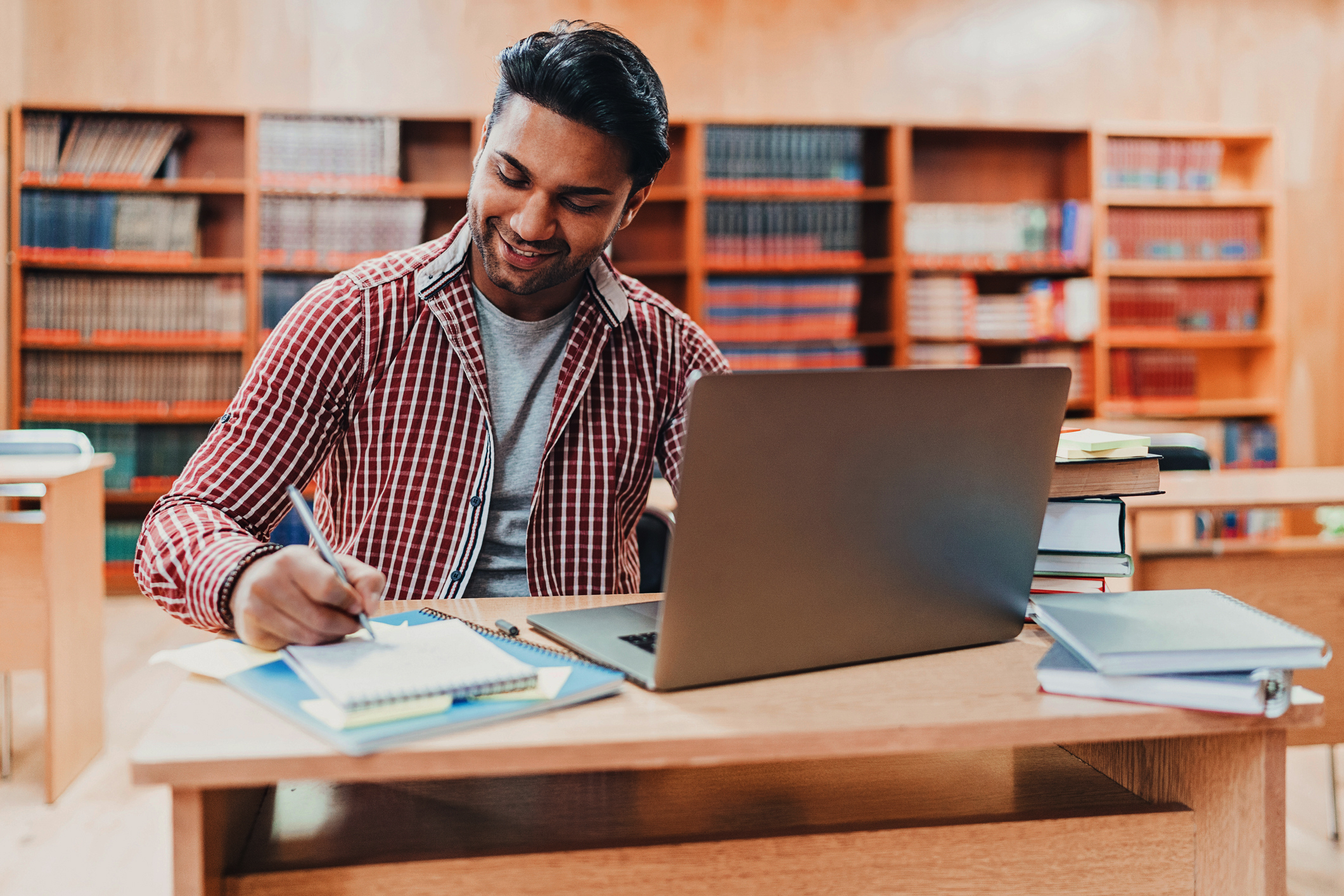 Male South Asian/Desi American college student studying in the library for his GRE exam. He has his laptop open on the library desk and is taking notes in a notebook.