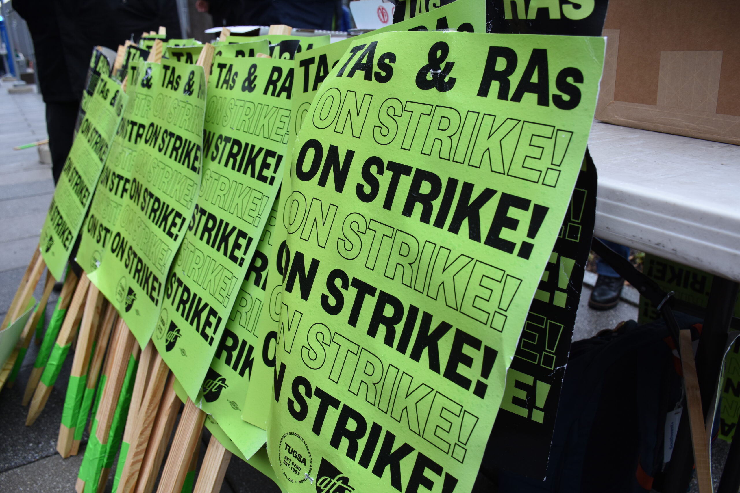 Printed student strike signs resting against a sidewalk bench on the Temple University campus. The signs read, "TAs and RAs On Strike!"