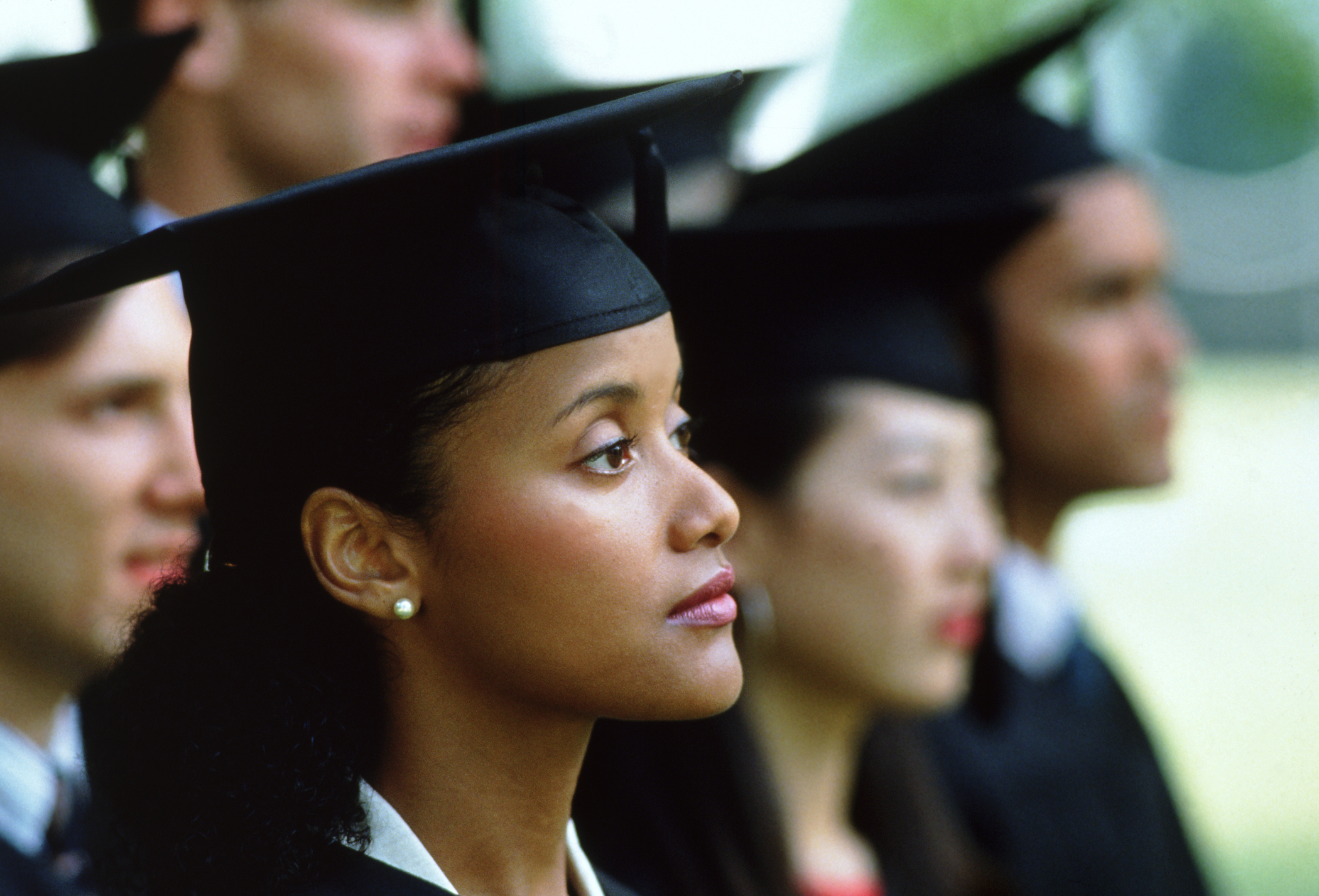 Black female graduate dressed in a cap and gown listens intently to a speaker at her college's commencement ceremony.