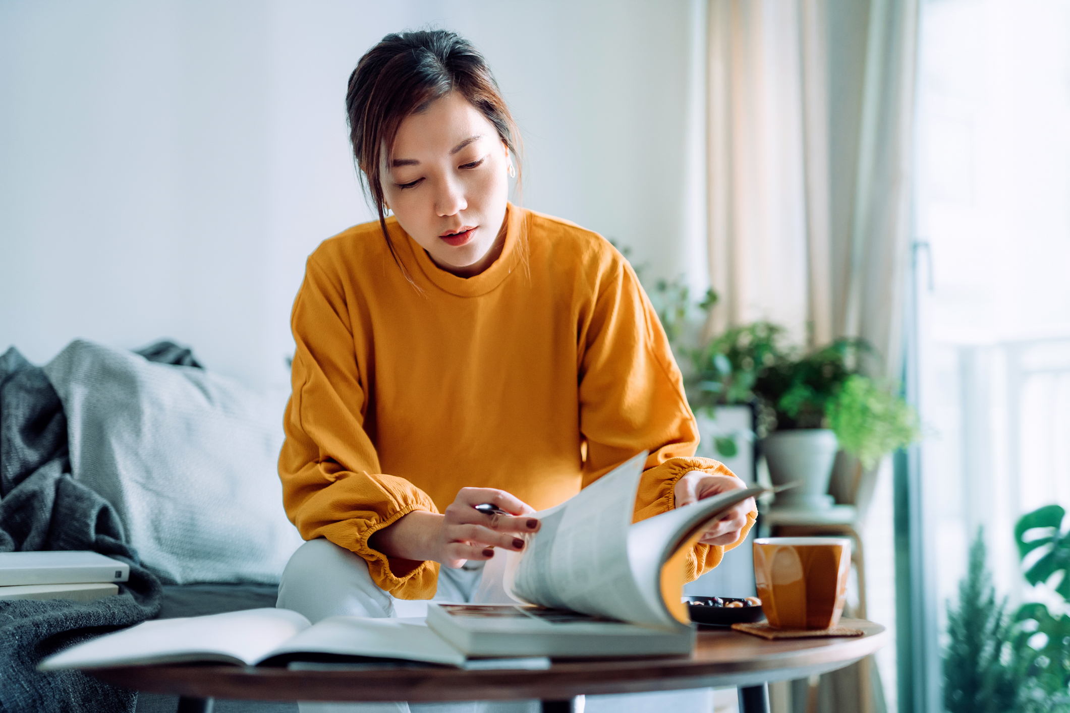 Female Asian American college student sitting at home on her living room couch. She is concentrating on studying for the LSAT exam. She is reading from an exam practice book and taking notes.