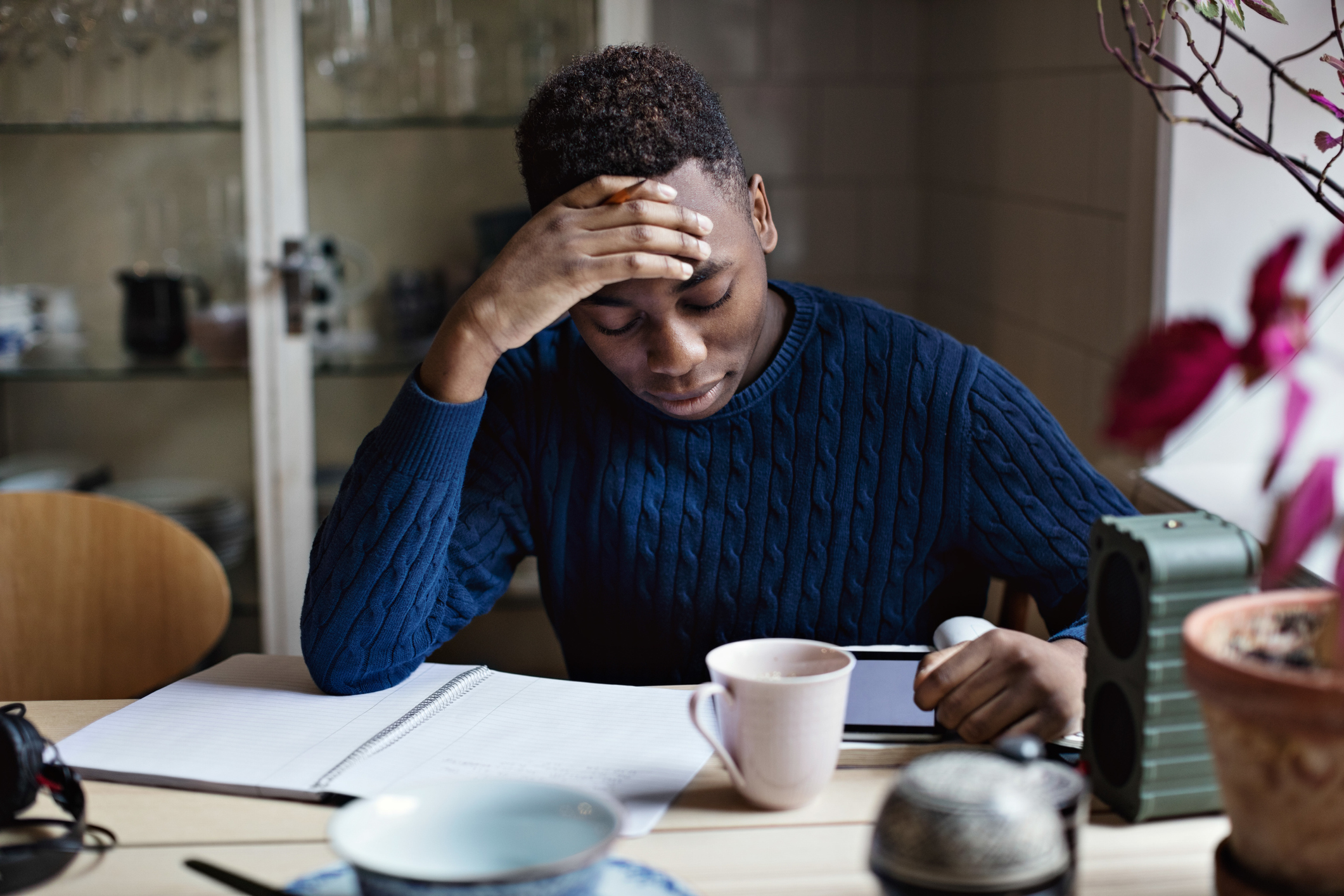 A Black male college student sitting at a desk in his living room at home. He is looking at reading materials on his tablet device and has a notebook open. He is resting his forehead on his right hand, and has a tired, overwhelmed look on his face.
