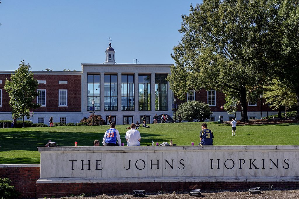 A view from Charles street of the grassy "Beach" and Milton S Eisenhower Library of the Johns Hopkins University; three male students, their backs facing the camera, sit on top of the marble university sign, other students sunbathing and playing catch on the grassy hill; the clock tower of Gilman Hall, a humanities building, peeks above the top of the library; Baltimore, Maryland, March, 2014. Courtesy Eric Chen. (Photo by JHU Sheridan Libraries/Gado/Getty Images).