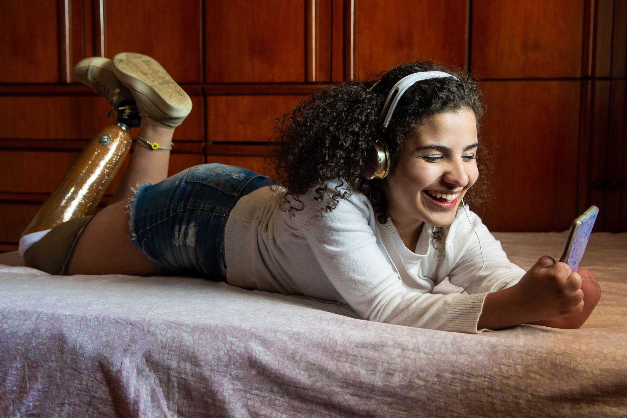 Female college student with prosthetic leg and symbrachydactyly hanging out on her bed in her dorm room. She's smiling while listening to music on her cellphone.