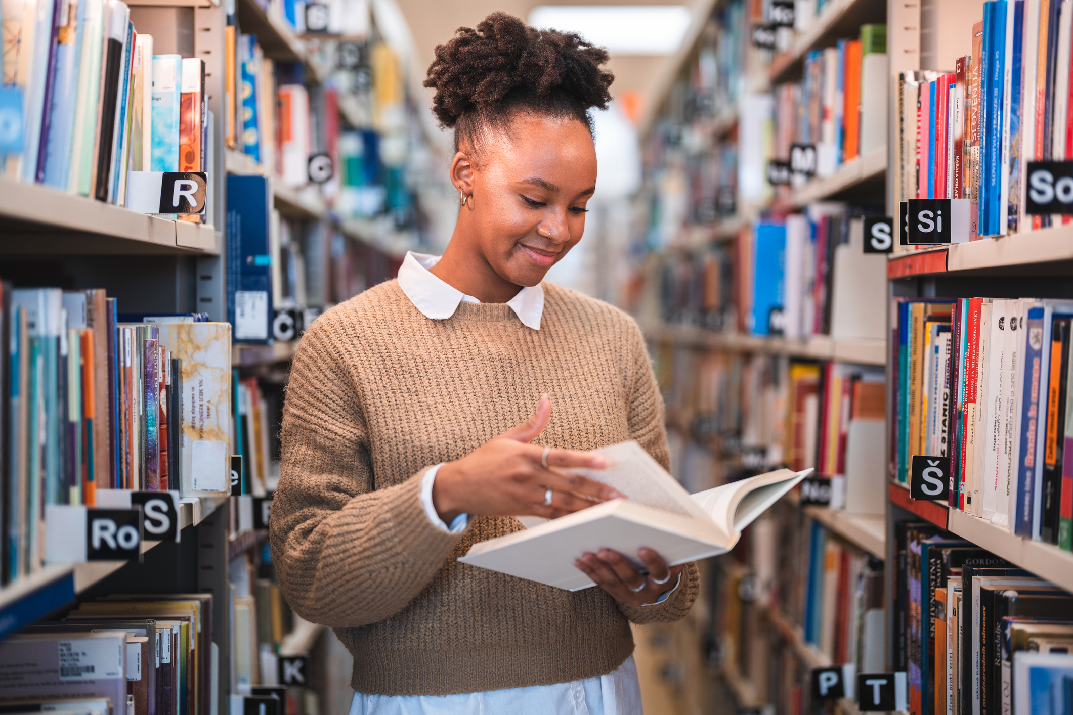 Latina college student standing in an aisle in a university library. She is smiling while reading a textbook for one of her courses.