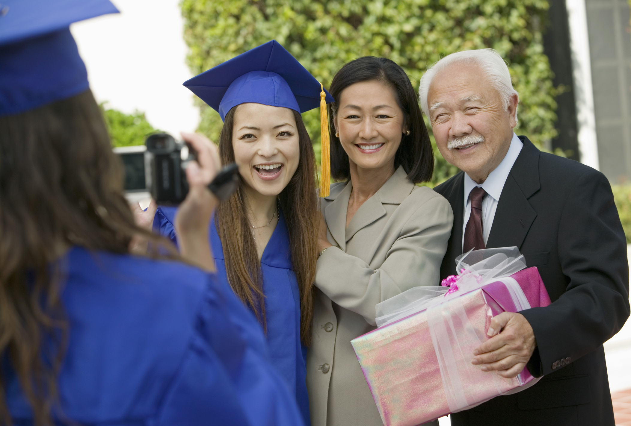 A first-generation Asian American female student poses with her mother and grandfather at her graduation ceremony. She is wearing a blue cap and gown. A friend is videotaping her and her relatives in celebration of her achievements.