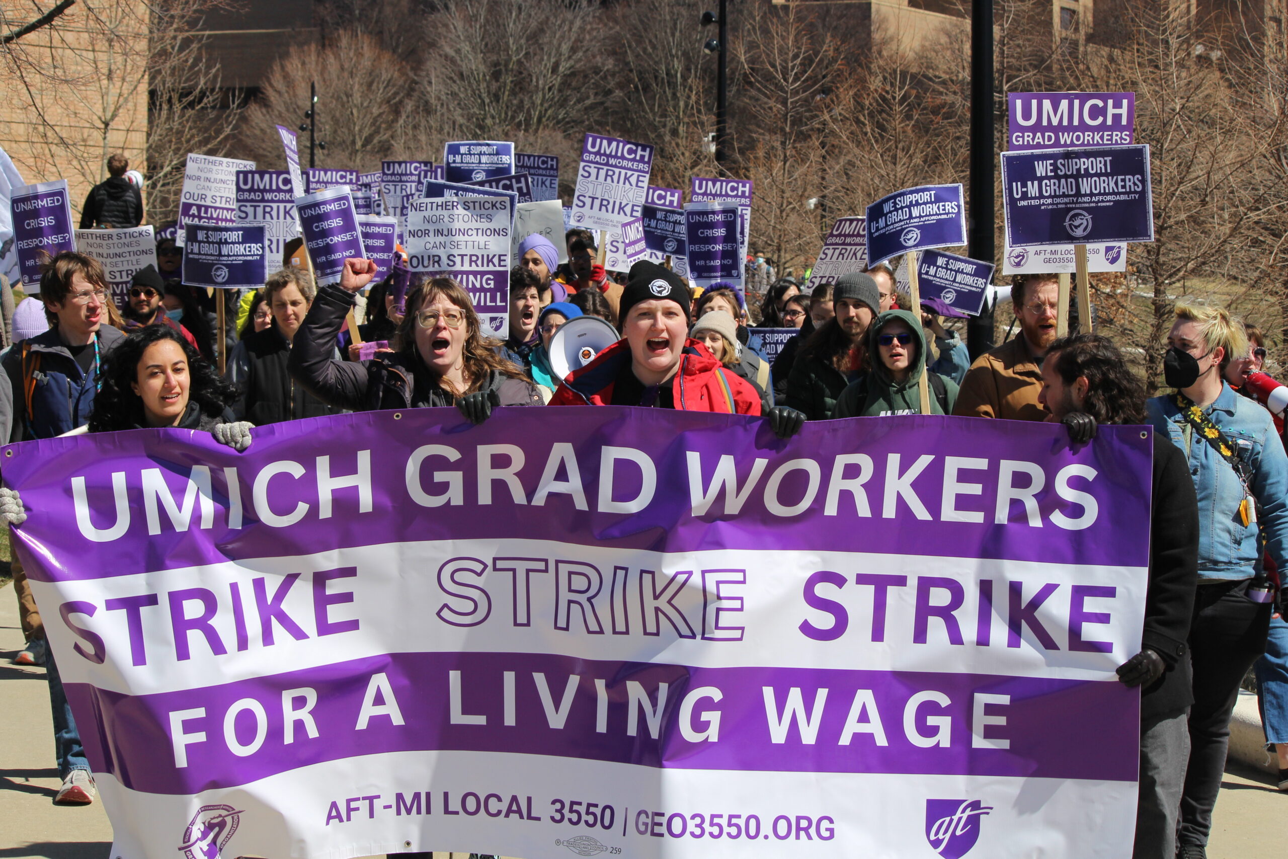 A group of University of Michigan students on strike. they are standing outside holding a large purple sign that says: UMich Grad workers strike for a living wage.