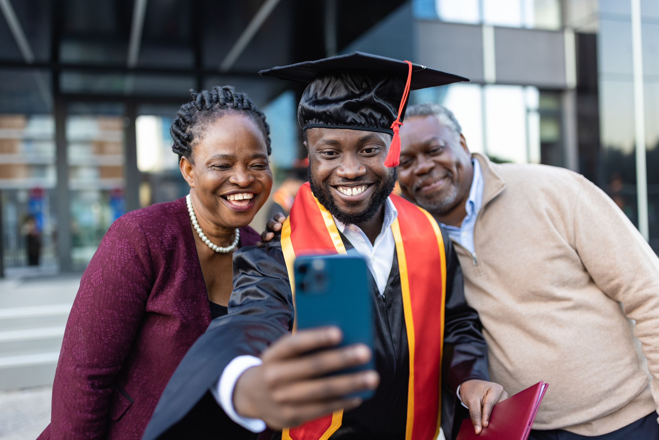 Excited Black male college graduate taking selfie photos with his proud parents after his graduation ceremony. He is wearing a black cap and gown and holding his degree in his left hand. He is using his right hand to take photos with his cell phone.