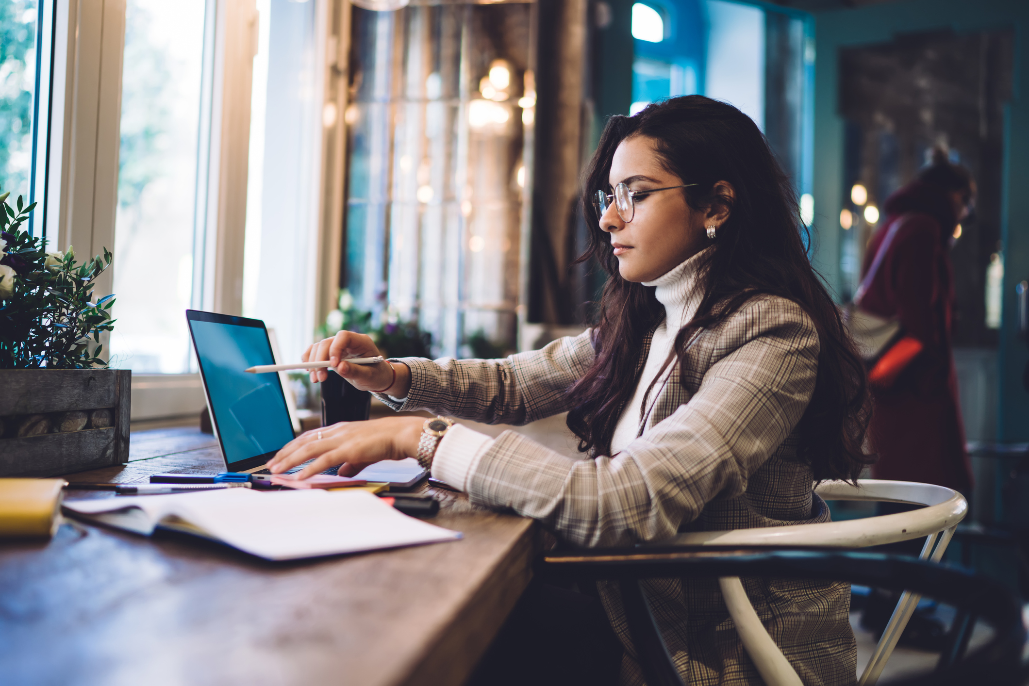 Female law student wearing glasses sitting at a table in a cafe. She is writing notes on a digital tablet. She is studying from several open textbooks.