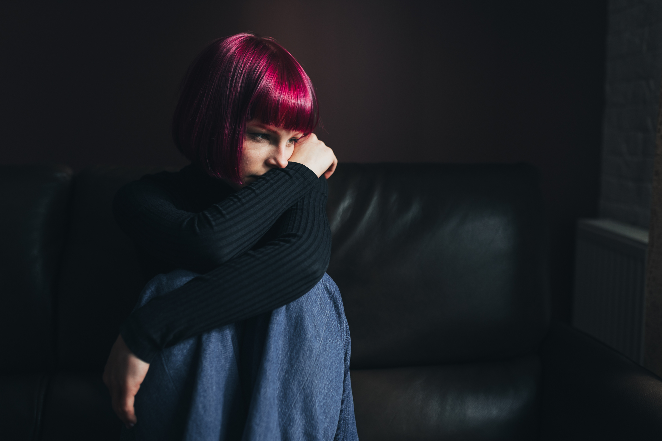 Sad female college student sitting alone on her living room couch with the lights off. She is hugging her knees to her chest and staring wistfully outside the window.