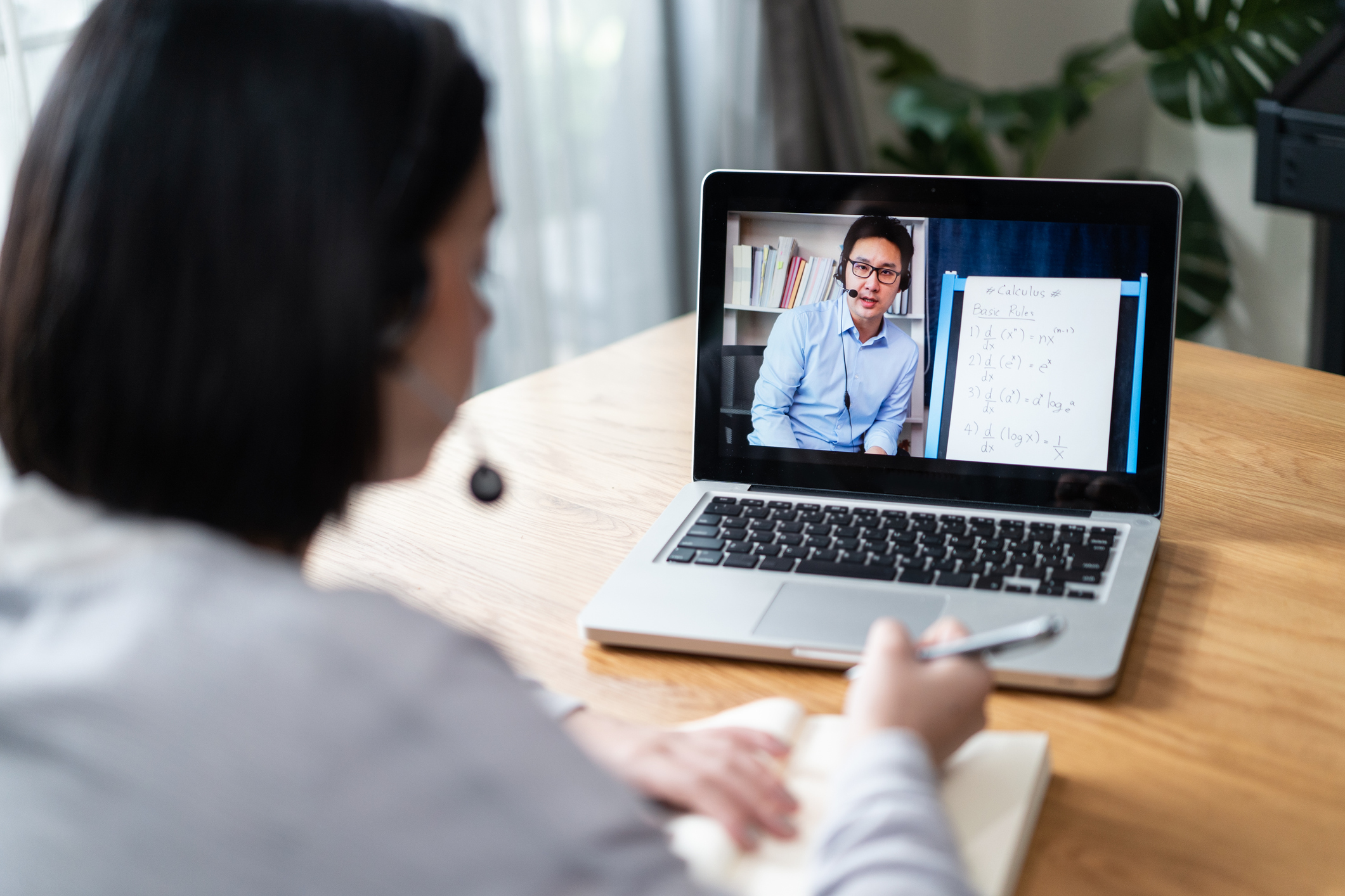 Student sitting in her home living room with her laptop open is taking an online calculus class for college. She is watching her instructor on her laptop screen demonstrating calculus equations on the whiteboard behind him.