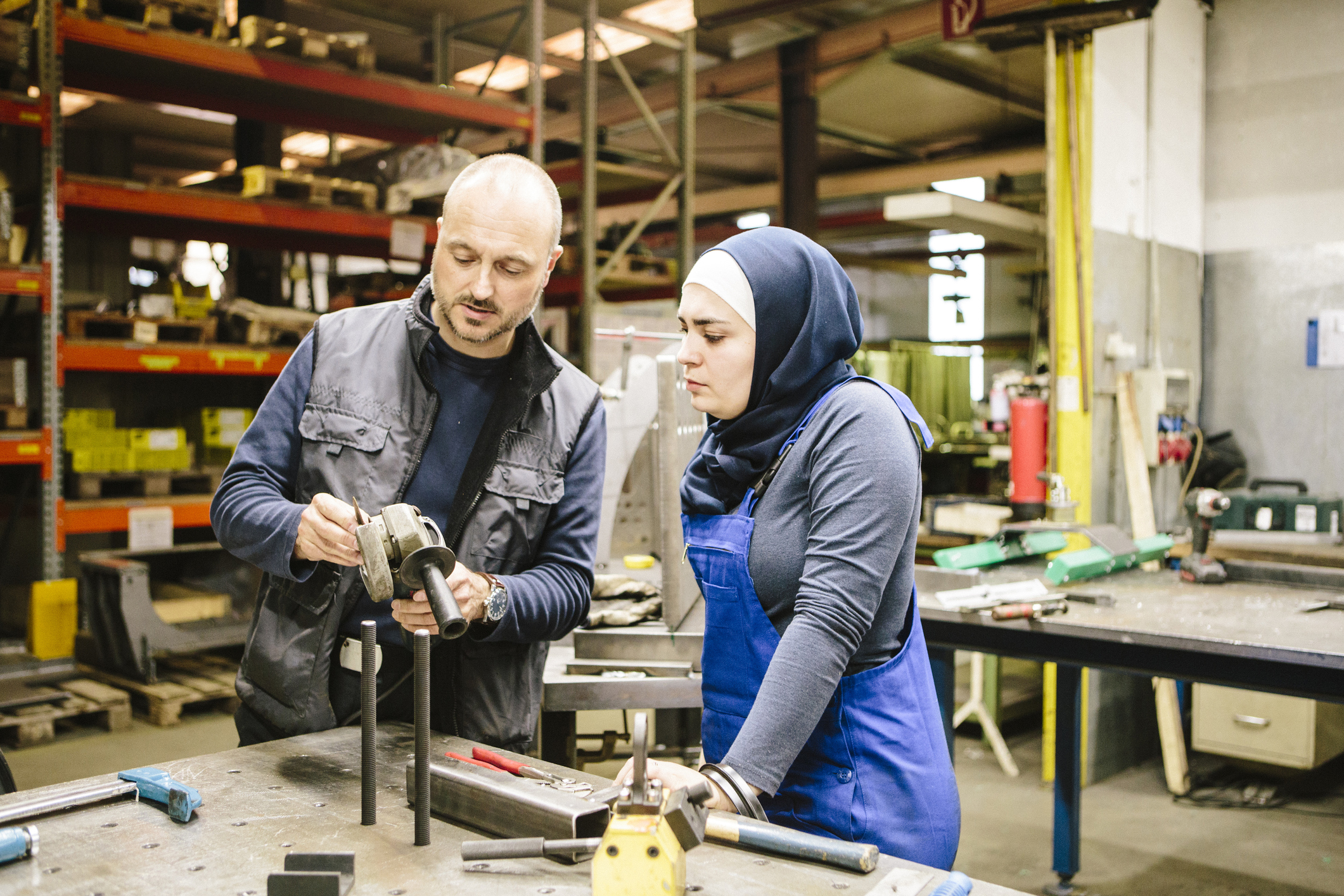 A male woodworking construction teacher instructing a female Muslim student on how to use a wood grinder. They are standing in a workshop on campus surrounded by woodworking machinery.