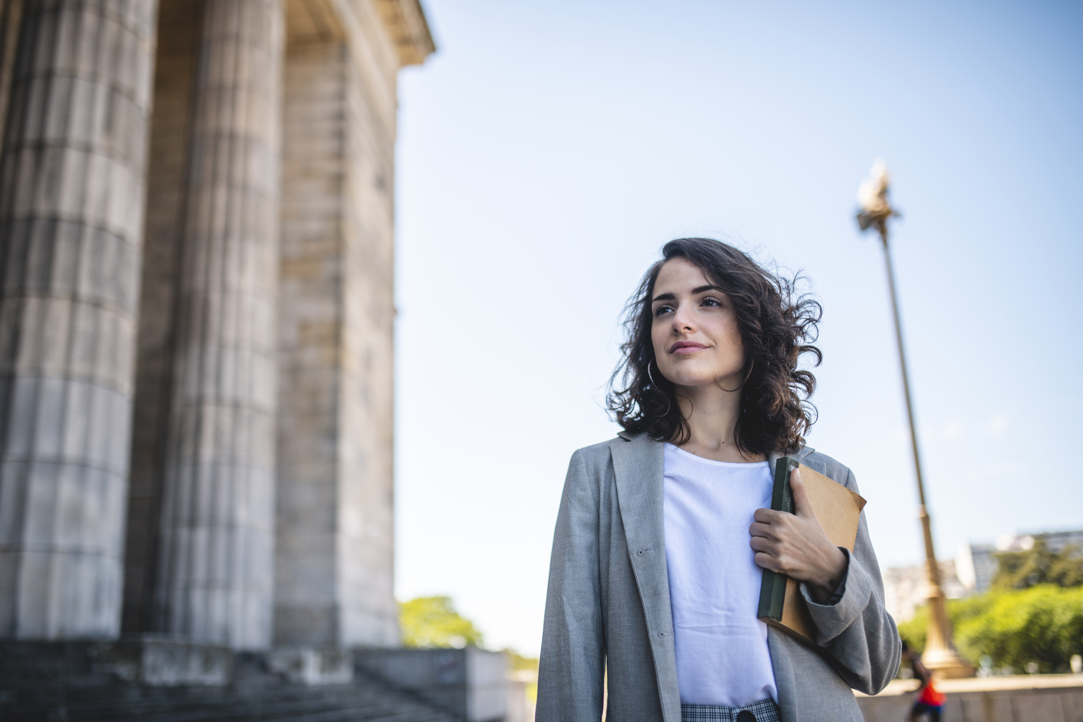 Female law student in her late 20s standing with a textbook at the entrance to a Neo-Classical university building on campus.