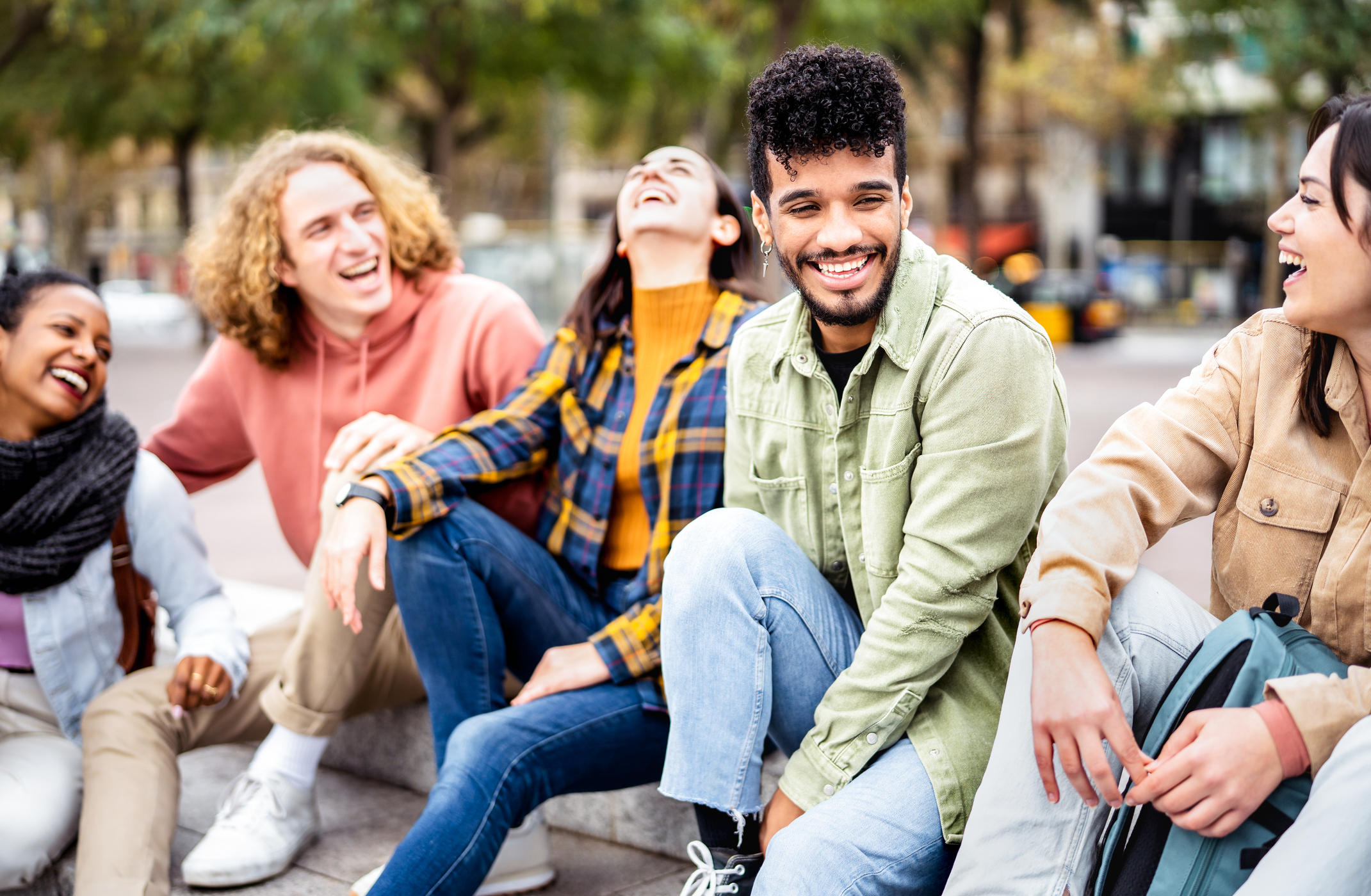 A multicultural group of male and female college students hanging out in a city park after class. They are sitting on a park bench, laughing over a shared joke with each other.