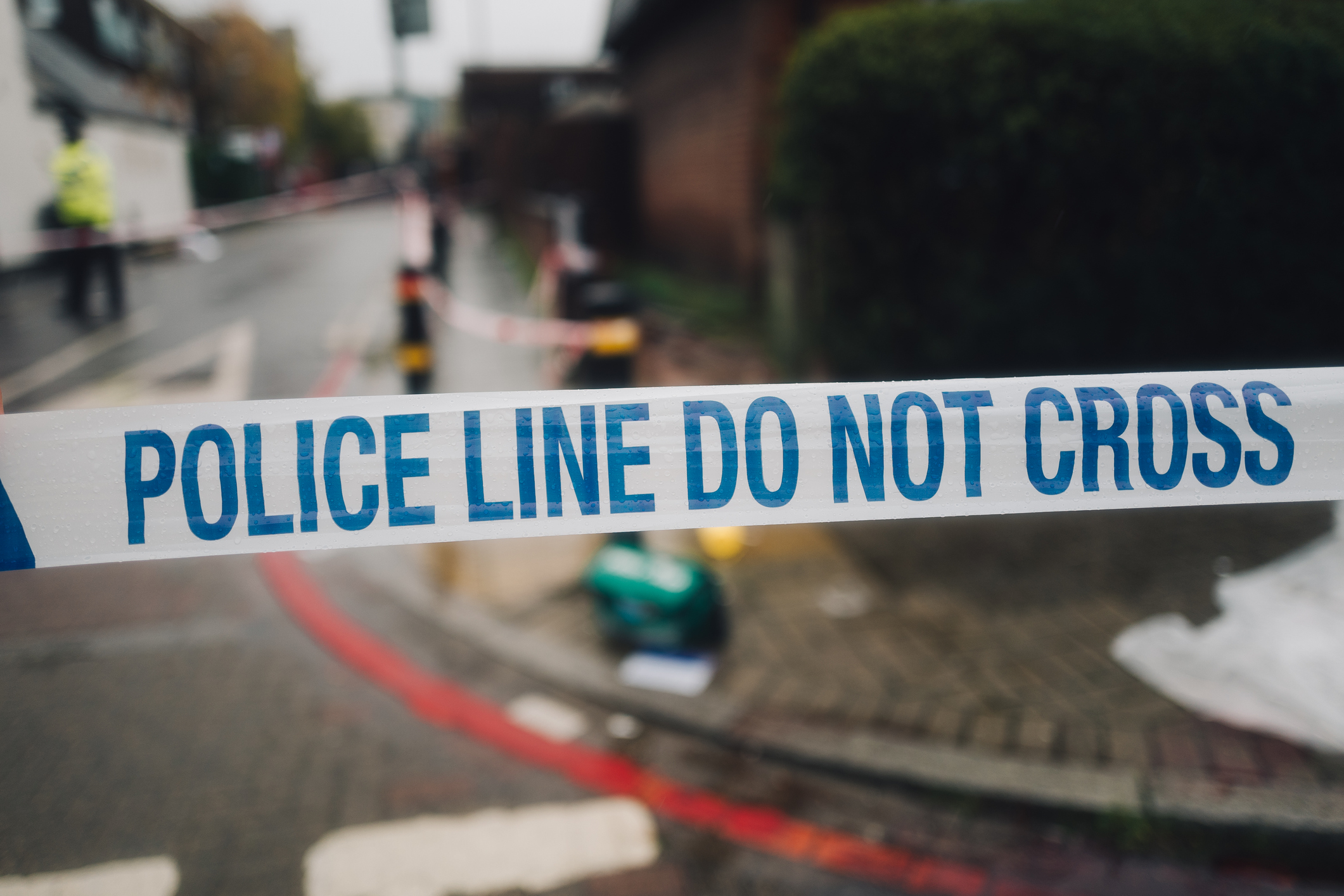 White and blue police cordon tape blocks the way to a crime scene on the street. Cops can be seen in the background walking around the scene during their investigation.