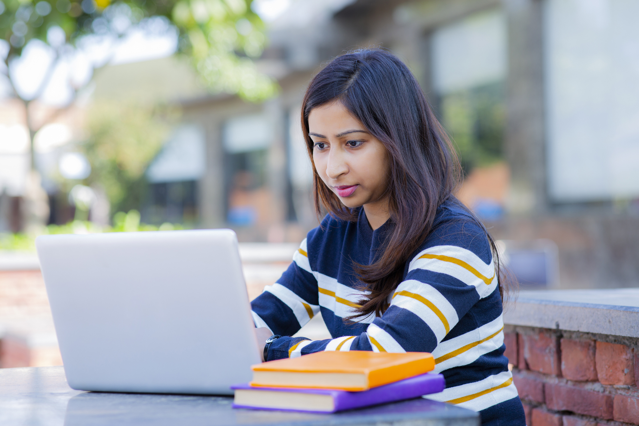 Female South Asian American student sitting at a table in an outdoor park on campus. She has a focused look on her face as she's typing on her laptop.