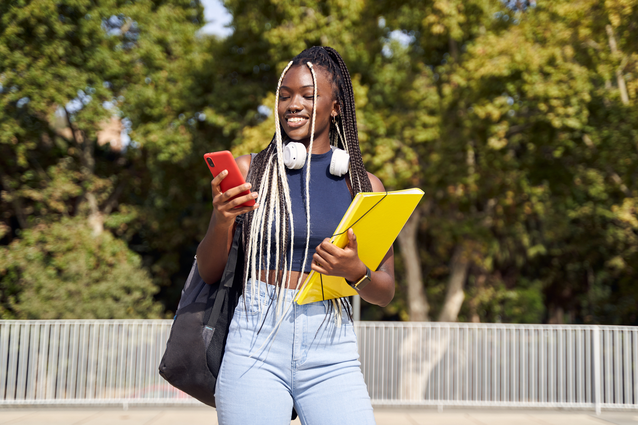 Cheerful young Black female college student standing outdoors in the city with backpack and folders in hand while using her mobile phone.