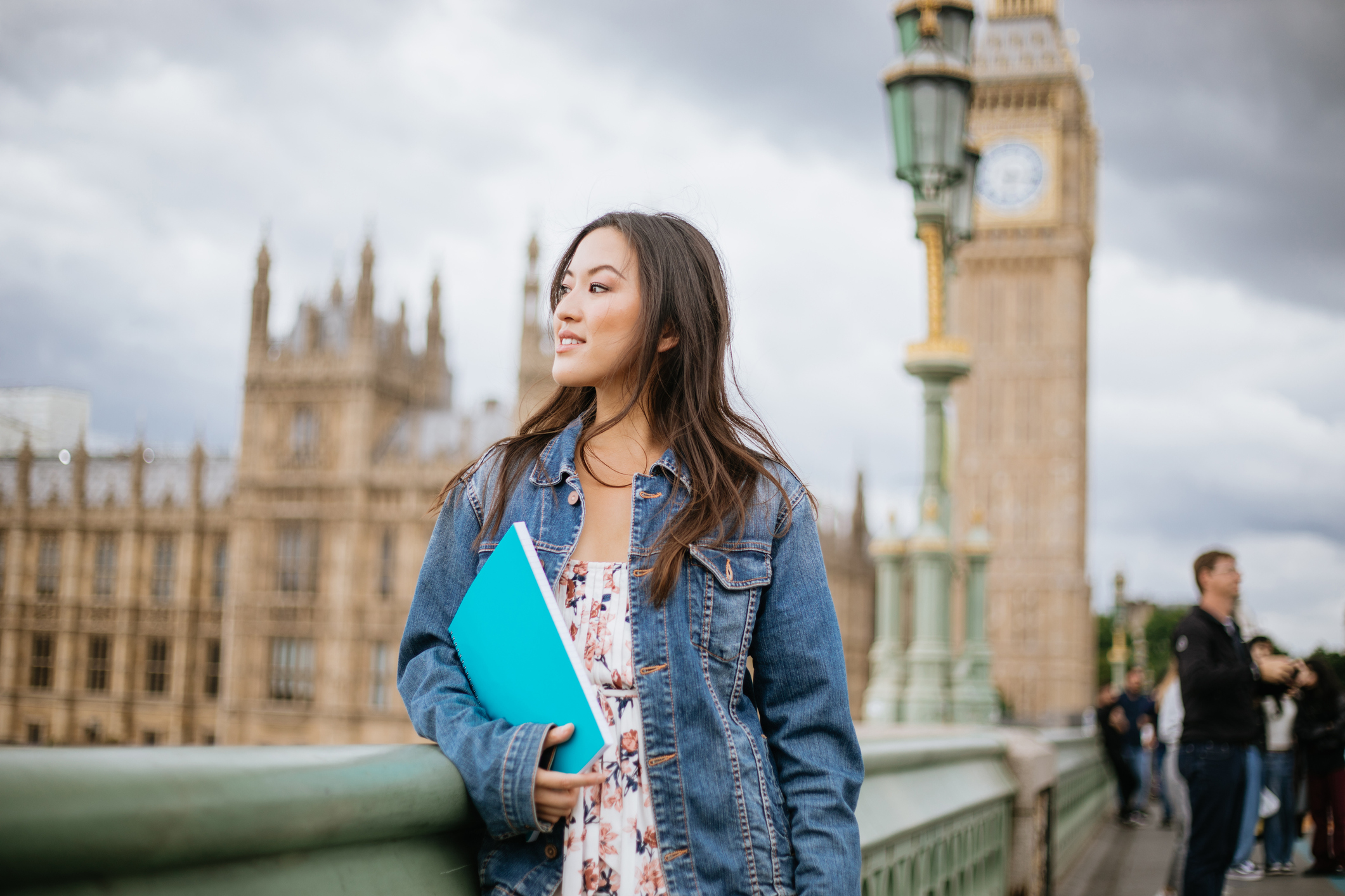 Asian American female college student standing on the Westminster Bridge in London, UK. She is holding a notebook. Big Ben can be seen in the background.