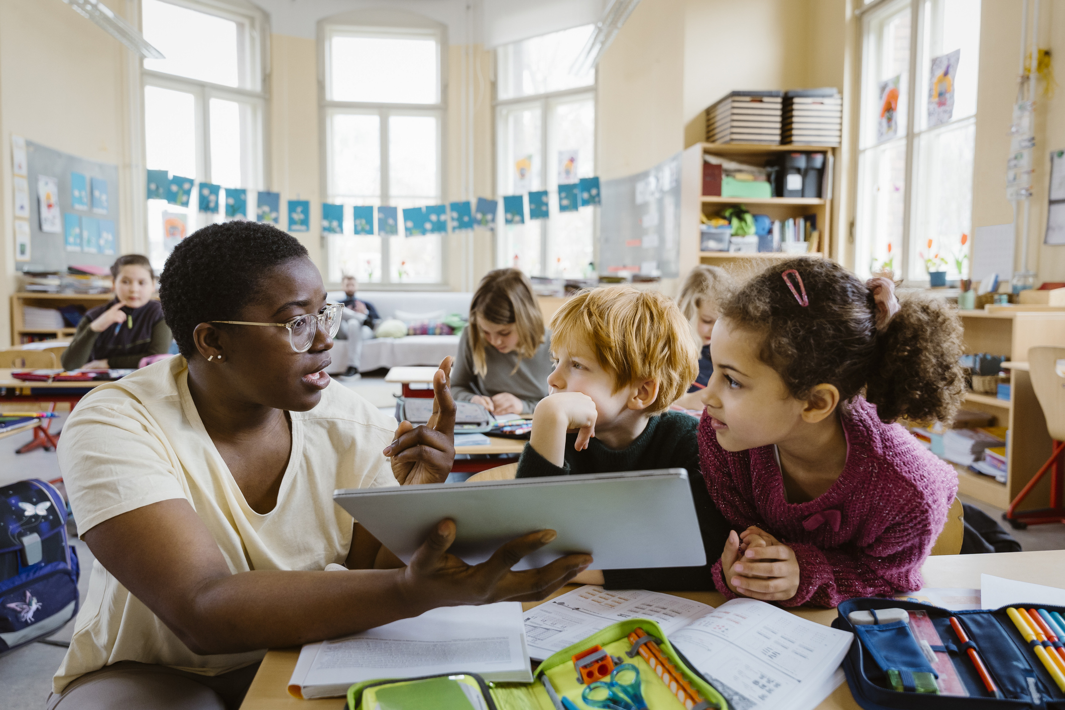 Black female teacher holding a tablet device to help explain a concept to an elementary-aged schoolboy and schoolgirl at their classroom desks.