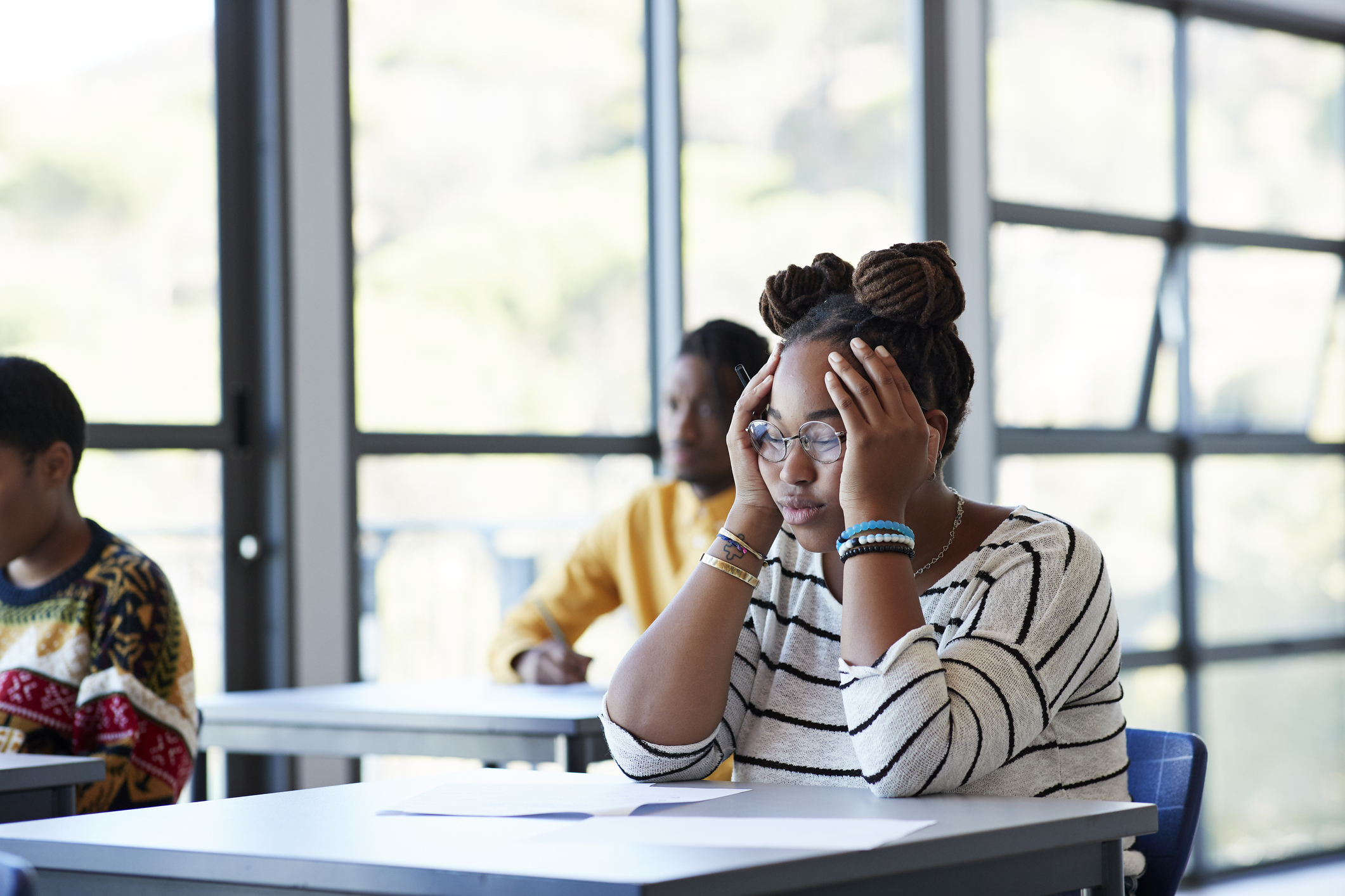 Worried young female student sitting with head in hands at desk during exam in community college classroom.