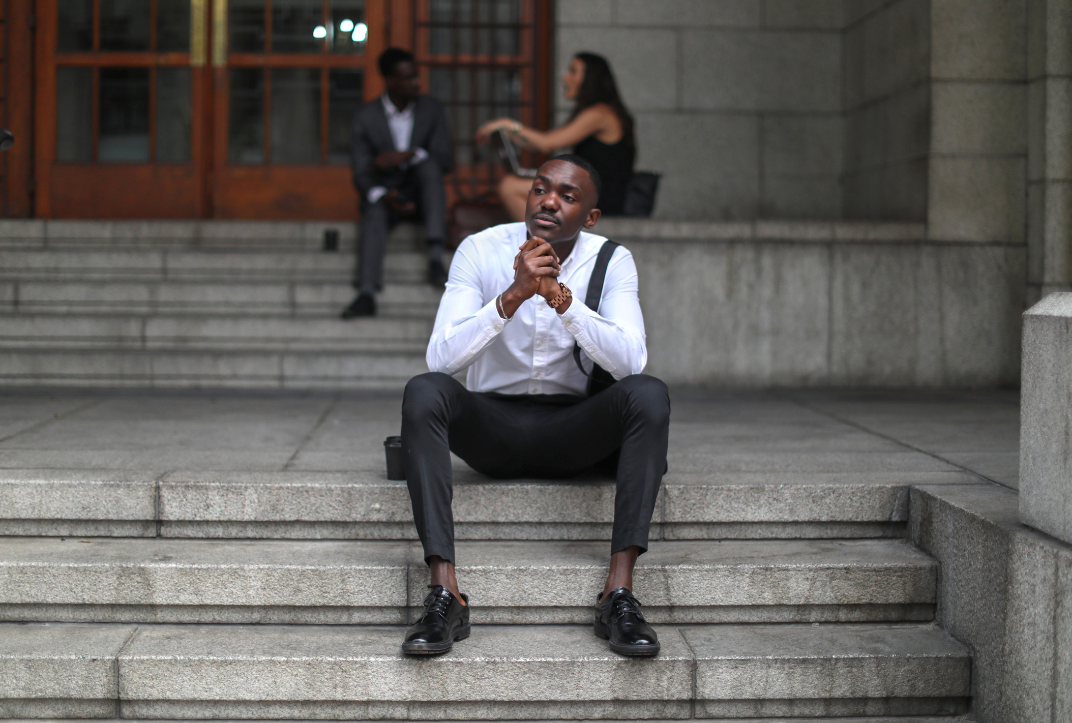 Black male lawyer sitting on steps. He is dressed in a business suit, and is looking offscreen with a thoughtful, serious look on his face.