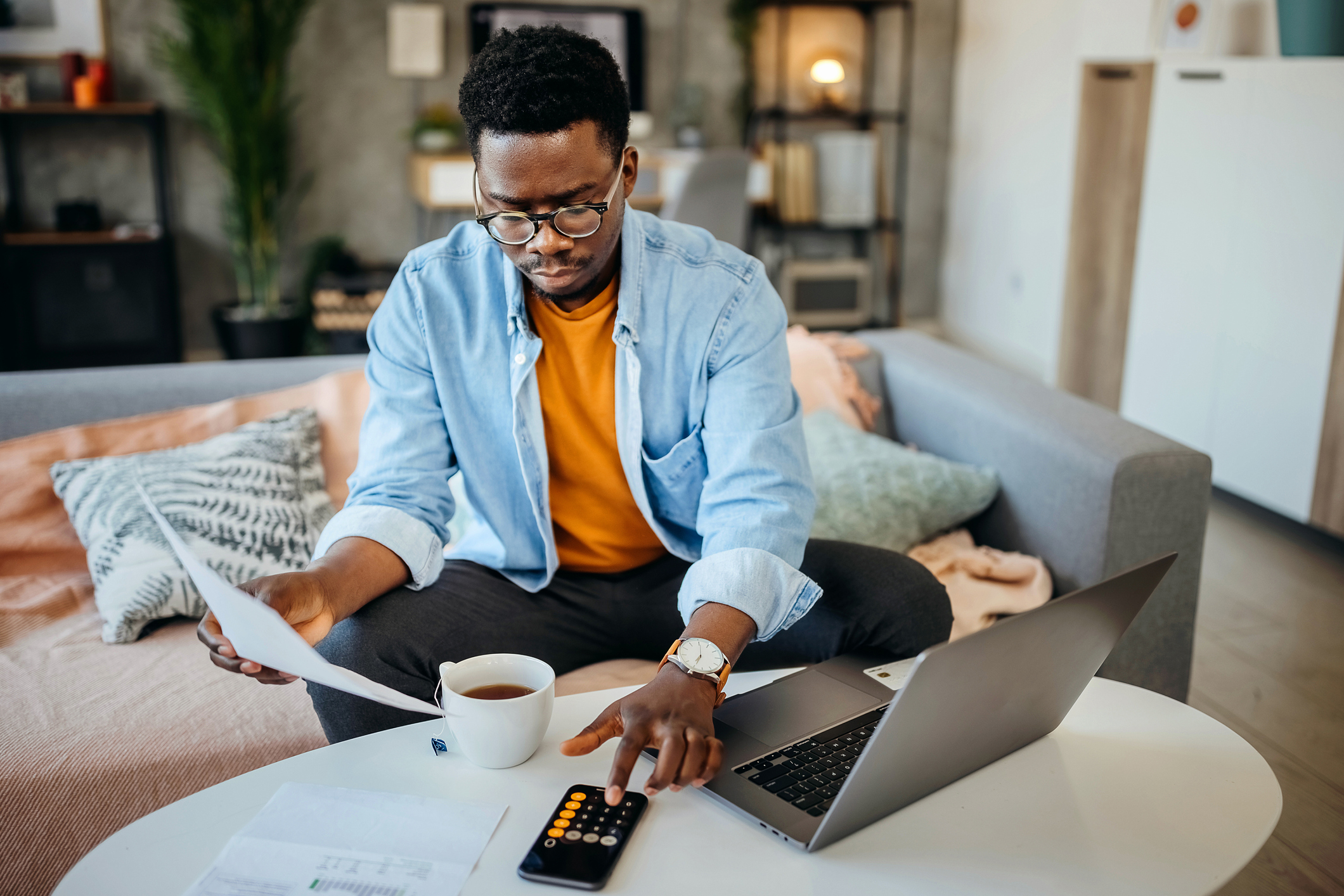 Black male college student sitting on a couch in his living room. He has his laptop open on the coffee table, and is holding a paper bill in his right hand. He is typing numbers on a calculator to check the bill amount on his student loan bill.