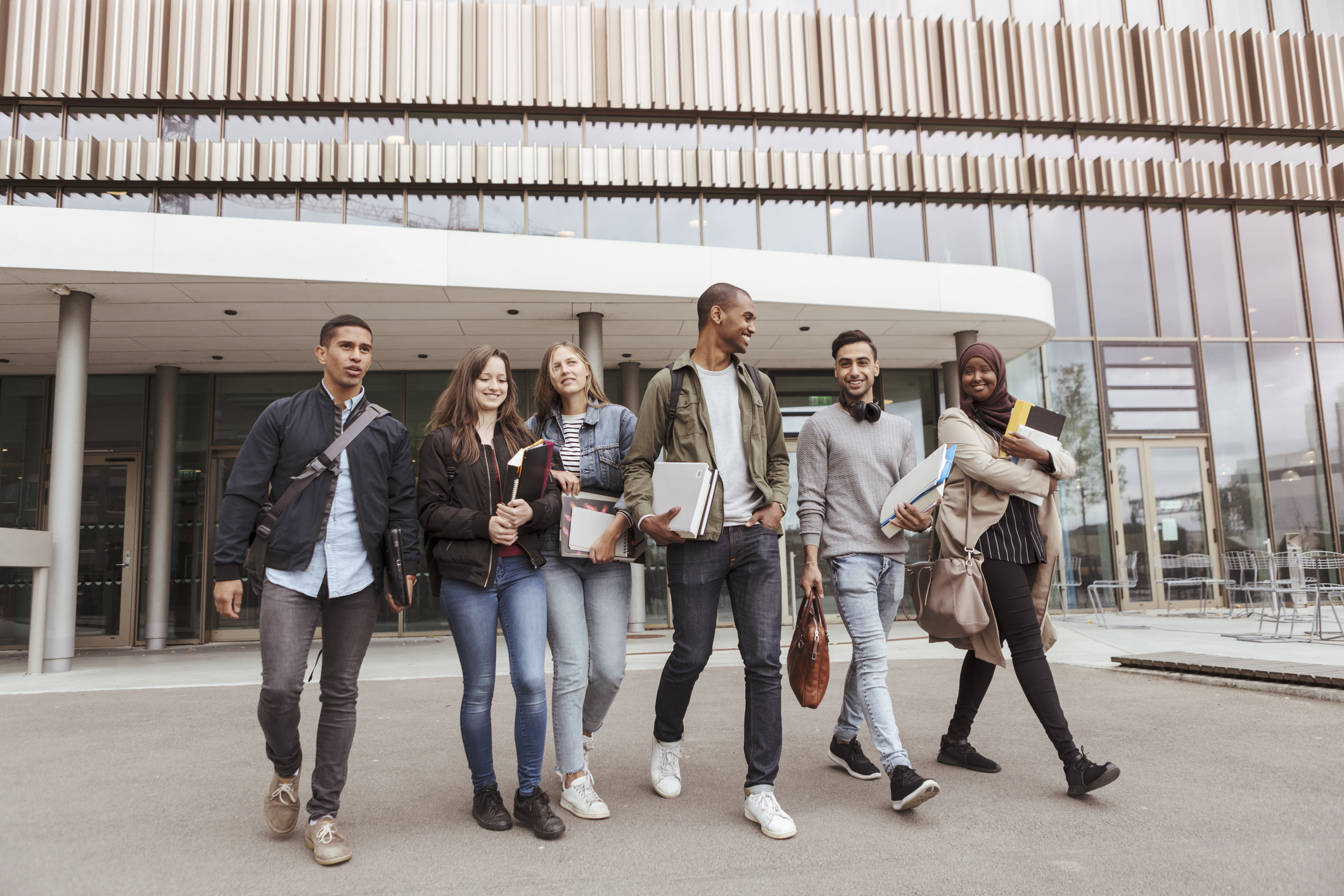 A diverse group of 6 college friends walking together on campus. They are walking out of a university building getting ready to hang out.