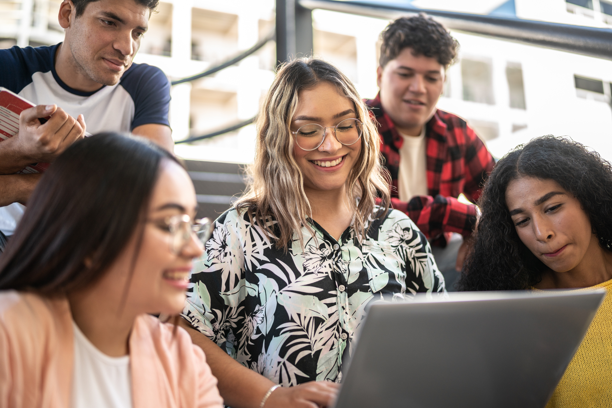 A Latina college student sits with her friends in a university classroom. She is smiling while showing them something on her laptop.