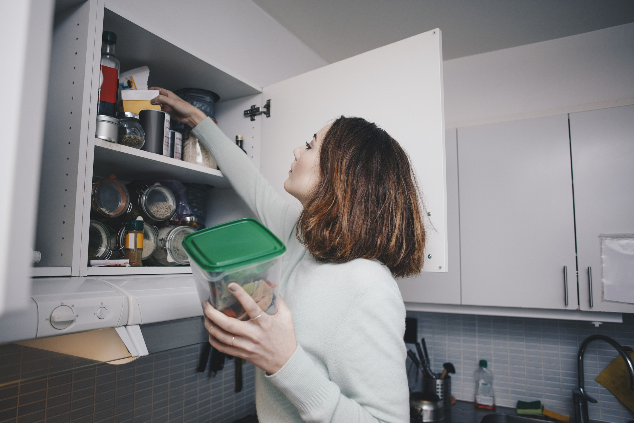 Female college student searching for ingredients in her apartment kitchen shelves.