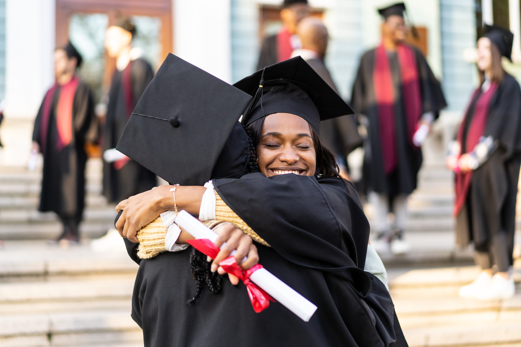 Two Black female college students are hug each other after receiving their diplomas on graduation day. They are wearing black caps and gowns and smiling.