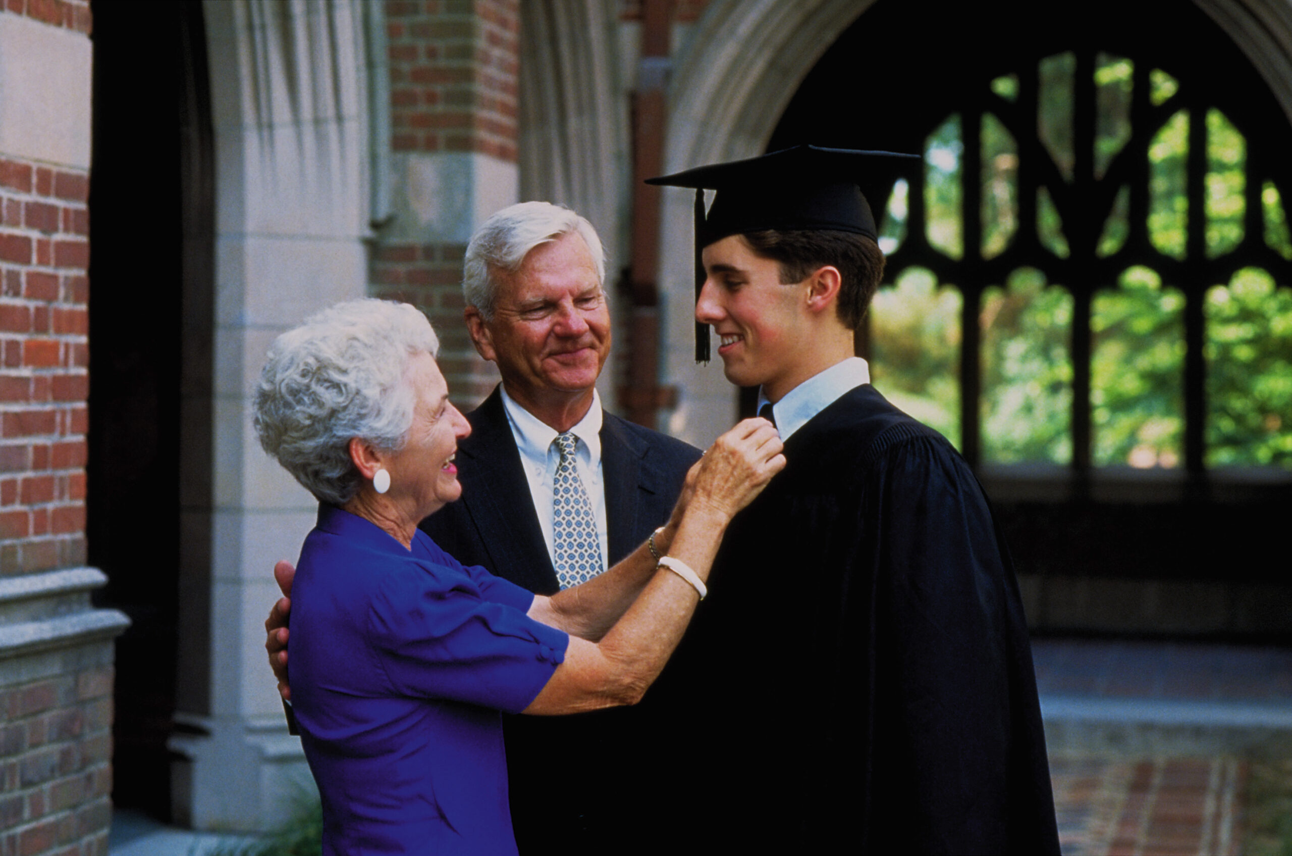 A Caucasian male college student dressed in a black cap and gown celebrates with his grandparents on his graduation day. His grandmother is adjusting his tie, while his grandfather gives him a congratulatory pat on the back.