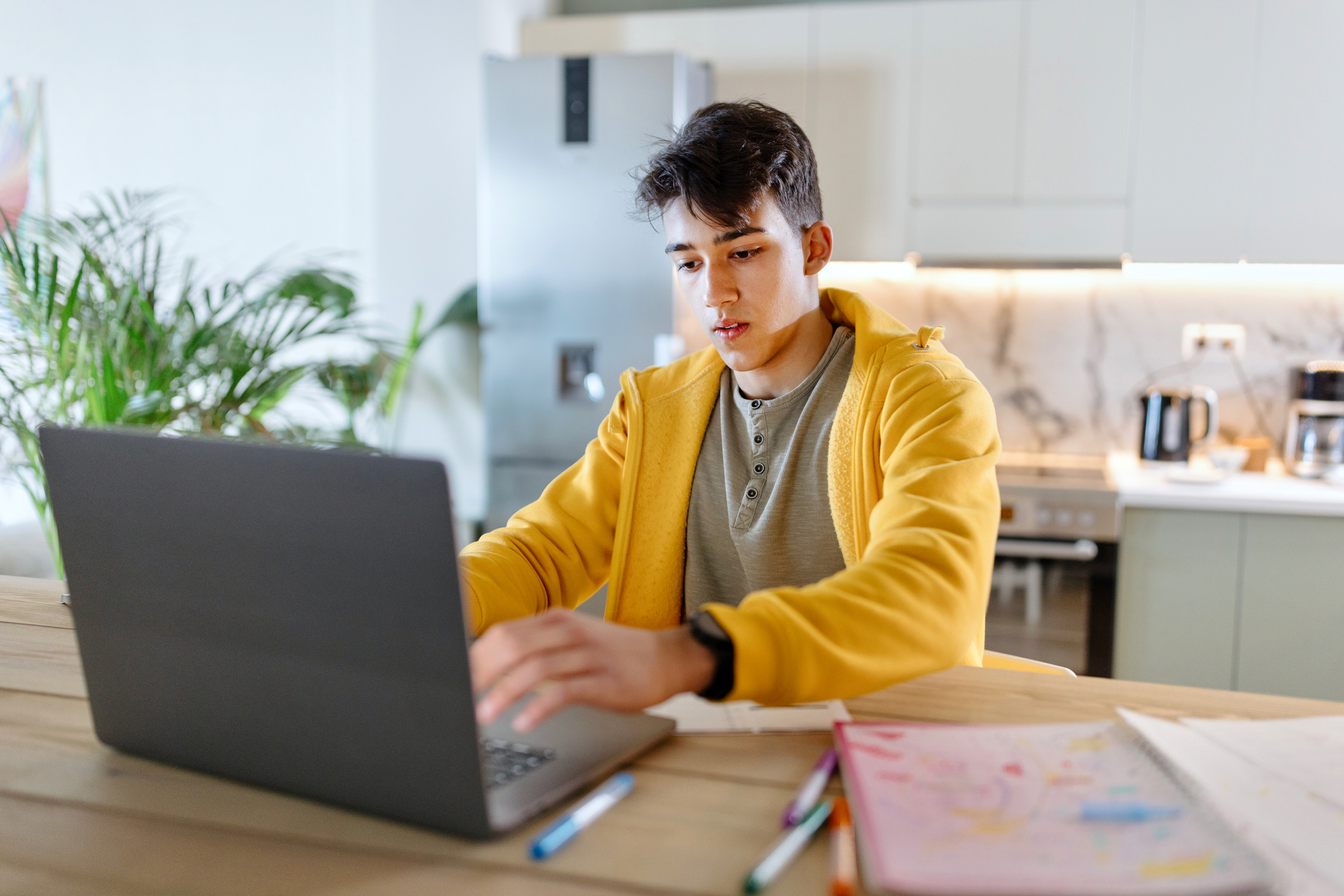 Male high school student sitting at a dining table at home. He is typing his college essay on his laptop computer.