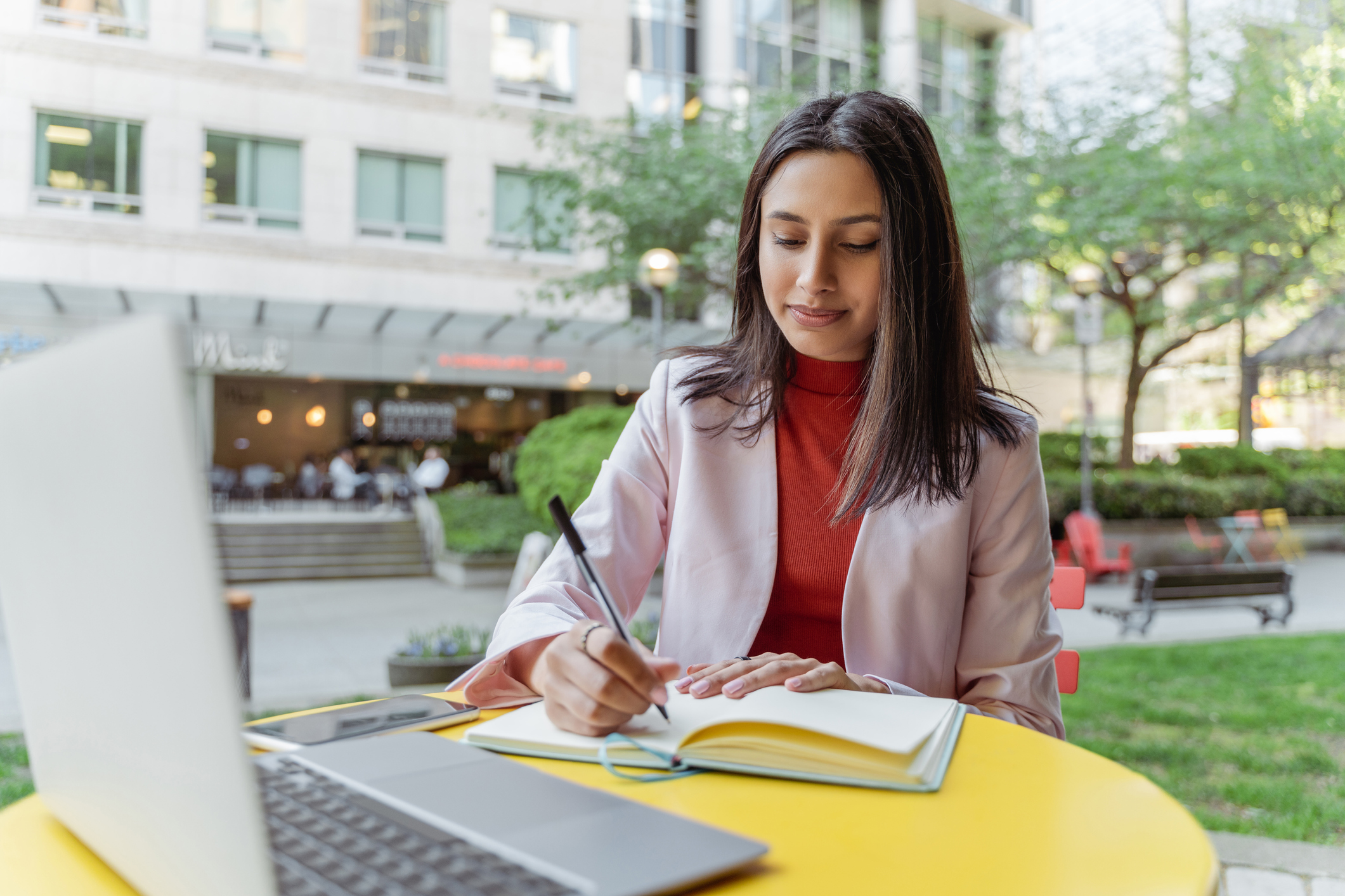 Female South Asian business student sitting at a table in an outdoor park. She is writing notes while referencing study materials on her laptop computer.