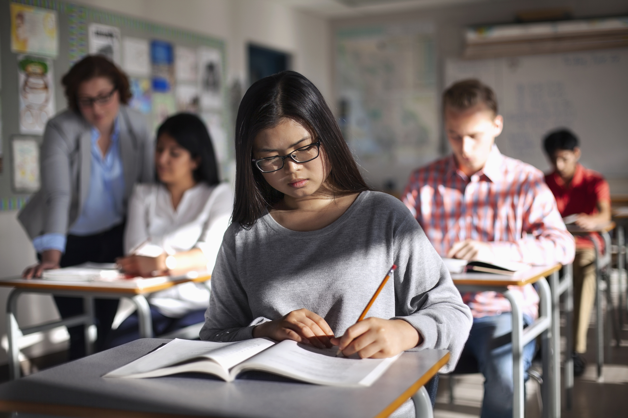 Asian American female student taking a standardized text in a high school classroom. She is sitting at her own desk, surrounded by several other students taking the exam.